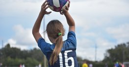 Ein Mädchen ist von hinten zu sehen. Ihr Pferdeschwanz ist dunkelblond und reicht bis über die Schulterblätter. Sie trägt ein dunkelblaues Trikot. Auf ihrem Rücken befindet sich die Nummer 16. Sie hält einen Fußball in beiden Händen über ihrem Kopf. Sie scheint ihn von der Seitenlinie aus ins Spielfeld werfen zu wollen. Im Hintergrund befinden sich verschwommen mehrere andere Fußballspielerinnen und -spieler. 