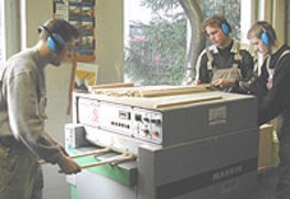 Drei Schüler arbeiten an einer Dickenhobelmaschine. Sie schieben Leisten in die Maschine bzw. nehmen sie ab. 