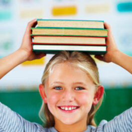 Ein lächelndes Mädchen balanciert (mit Unterstützung beider Hände) vier Bücher auf dem Kopf.