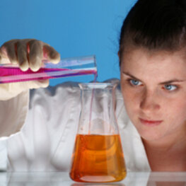 Ein Mädchen im Laborkittel schüttet eine Chemikalie aus einem Reagenzglas vorsichtig in einen Erlenmeyerkolben.