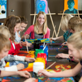 Um einen großen, rechteckigen Tisch sitzen mehrere Kinder mit Farbdosen in der Mitte, die verschiedene Bilder malen.