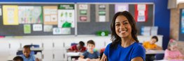 In einem Klassenraum steht ein junge, dunkelhäutige Lehrerin mit langen schwarzen Haaren. Im Hintergrund arbeiten einige Kinder an ihren Tischen.