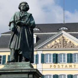 Eine Beethoven-Statue ist im Vordergrund links zu sehen. Sie steht vor dem alten Bonner Postamt, welches rechts im Hintergrund auszumachen ist.