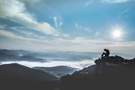 Ein Mensch sitzt allein auf einem Berg in weiter Landschaft.