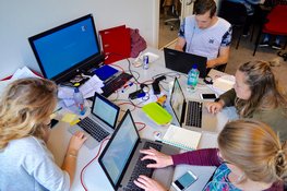 Gruppe von jungen Erwachenen, die an einem großen Tisch mit vielen Laptops sitzen.