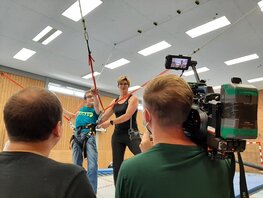 In einer Sporthalle filmt ein Kamerateam eine Lehrerin mit ihrem Schützling.