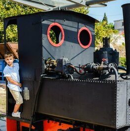 Auf einer kleinen, historischen Dampflok steht ein kleiner Junge im Zugführerhäuschen und schaut seitlich heraus.