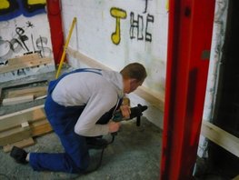 Ein Schüler bohrt mit dem Bohrhammer durch ein Brett in die Wand hinein, um danach das Brett mit einem Nageldübel zu befestigen.